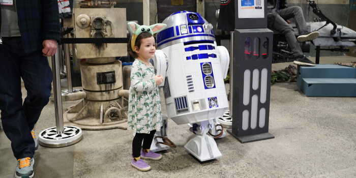 a little girl in a dinosaur dress, wearing a headband featuring Grogu's eyes and ears, poses excitedly with R2-D2 at the 501st booth.