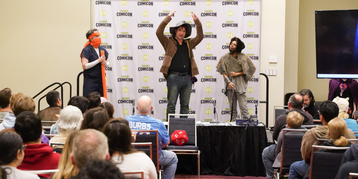A packed house for D+D live with Bad Dog Comedy. A D20 is on a chair at the front of the room, having landed on 5. The three performers are standing on stage. Far right is and far left are staring intently at the person in the middle, whose arms are raised in a spell casting.