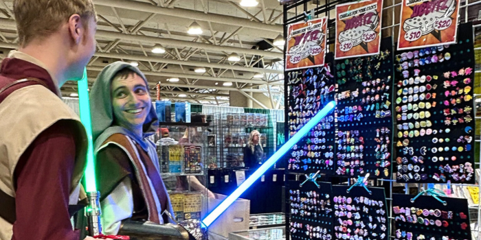 A pair, dressed as jedis, shop for croc jibbitz in artist alley. The one in the front smiles back to his friend while pointing at product with his illuminated saber