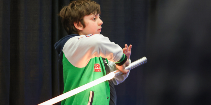 Child wearing a white and green jacket attends light saber training and poses with a white light saber.