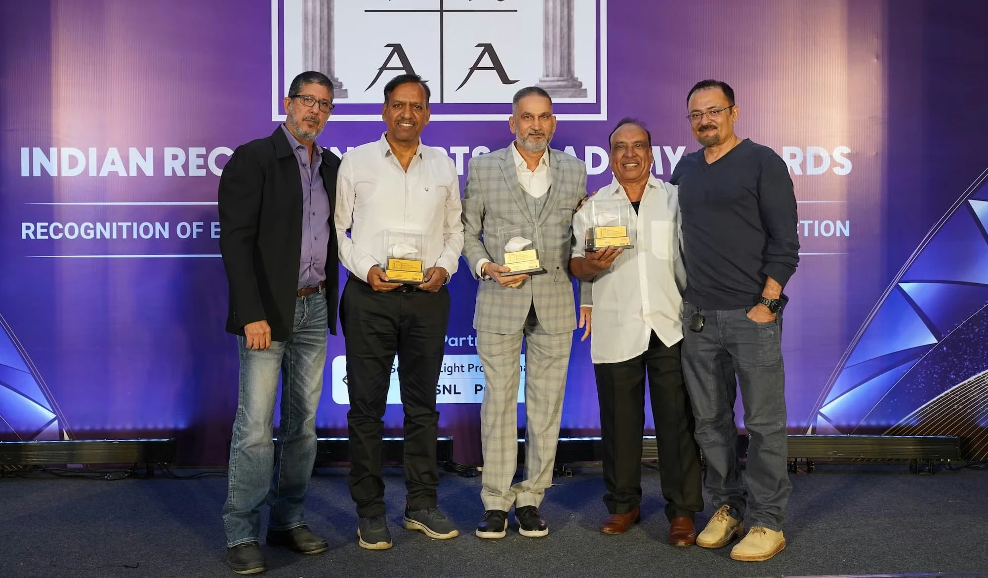 Seven people posing at the IRAA Palm Awards photo call with the event logo behind them, three pairs each holding a trophy, and the person on the far right simply posing for the photo.