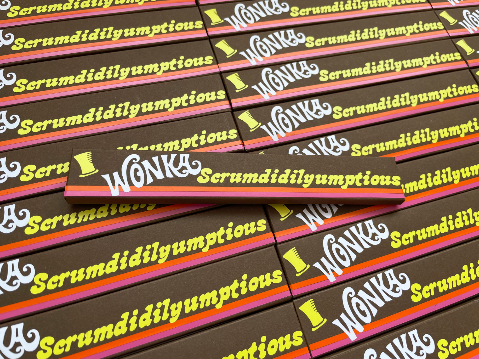 A background of angled Scrumdidilyumptious bars lays across the background, with one positioned straight across the top. The packaging is brown, with yellow text (excluding the white Wonka logo), featuring two stripes, one red, one pink.