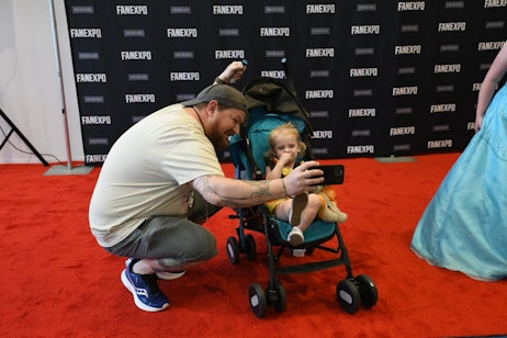 A dad excitedly takes a selfie with his young child, in stroller, on the cosplay red carpet with the FAN EXPO step and repeat in the background