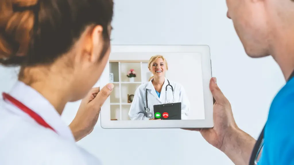 Profissionais de saúde de costas segurando um tablet durante uma chamada de telemedicina com uma médica sorridente na tela, representando o atendimento médico à distância do projeto TeleNordeste.