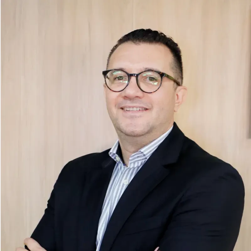 Adriano Ferreira, palestrante do Healthcare Innovation Show, homem sorrindo de óculos e camisa listrada com blazer.