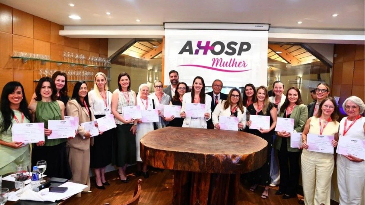 Grupo de profissionais, em sua maioria mulheres, posando lado a lado em um ambiente interno elegante, segurando certificados. Ao fundo, há um banner com a inscrição “AHOSP Mulher”. Os participantes usam crachás e trajes formais, sugerindo um evento corporativo ou cerimônia de reconhecimento. No centro da sala, há uma grande mesa redonda de madeira, e ao redor, estantes com taças e elementos decorativos.