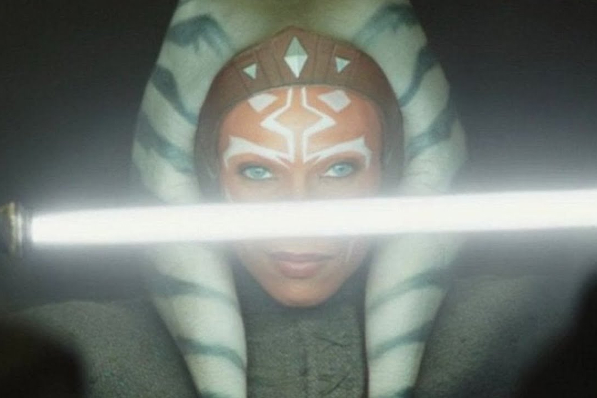 A close up of Ahsoka Tano's orange face with white and blue head piece holding an ignited white lightsaber infront of her