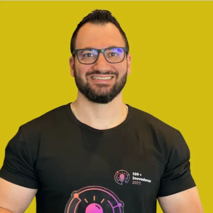 Alex Vieira, palestrante do Healthcare Innovation Show, sorri usando óculos e camiseta preta sobre fundo amarelo.