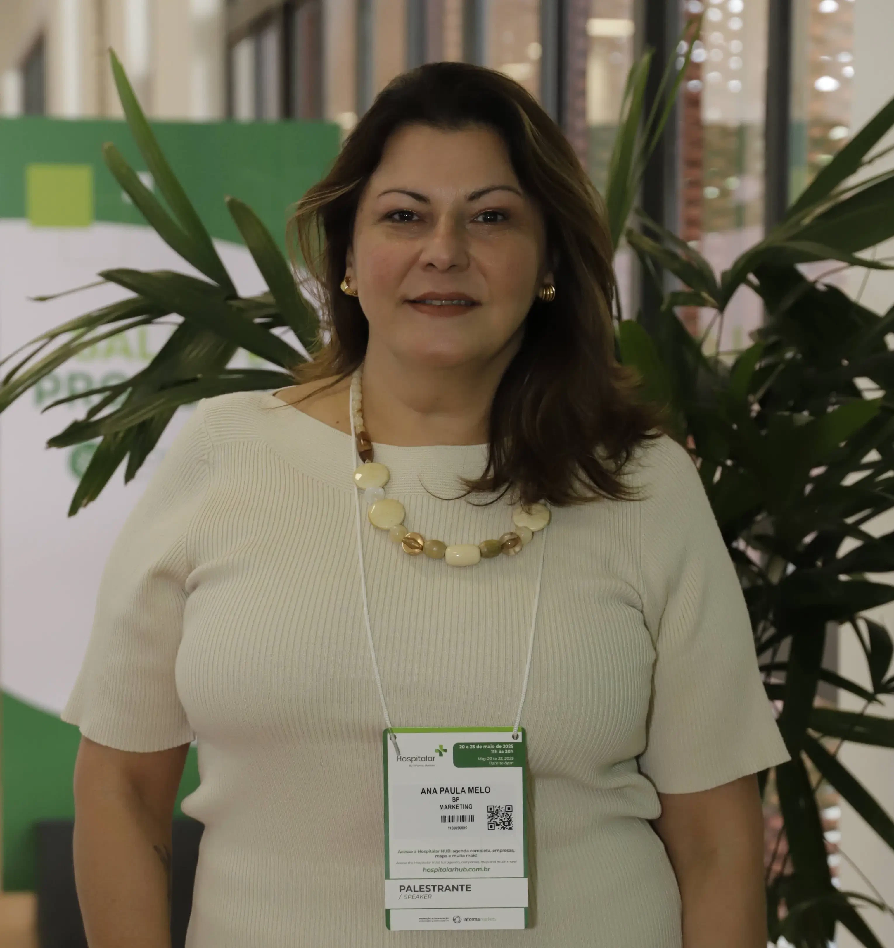 Ana Paula Melo, curadora e palestrante no CSC - Congresso de Supply Chain 2026, posa sorrindo em frente a uma planta ornamental. Ela utiliza crachá oficial da Hospitalar identificando sua atuação em Marketing e sua posição como palestrante.