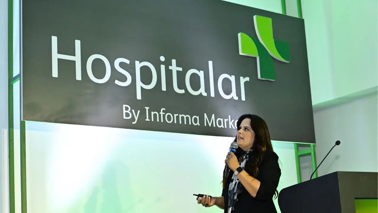 Palestrante apresenta conteúdo em evento da Hospitalar, com microfone em mãos e slide ao fundo com a marca Hospitalar by Informa Markets — conceito de congresso, inovação em saúde e networking no setor hospitalar.