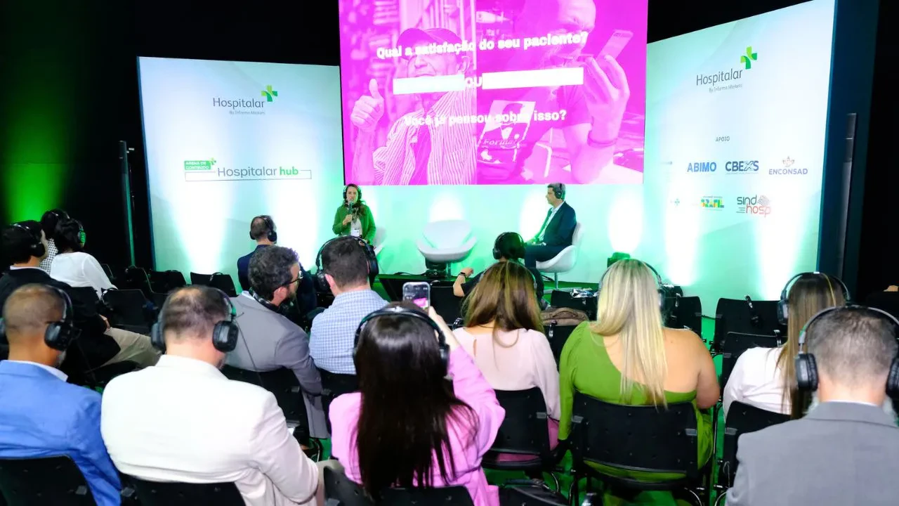 Público sentado em um evento corporativo de saúde, usando fones de ouvido para tradução simultânea, assiste a uma entrevista no palco com duas pessoas em poltronas. Ao fundo, há um telão com apresentação visual em tons de rosa e a marca “Hospitalar hub” exibida nas laterais do palco iluminado em verde.
