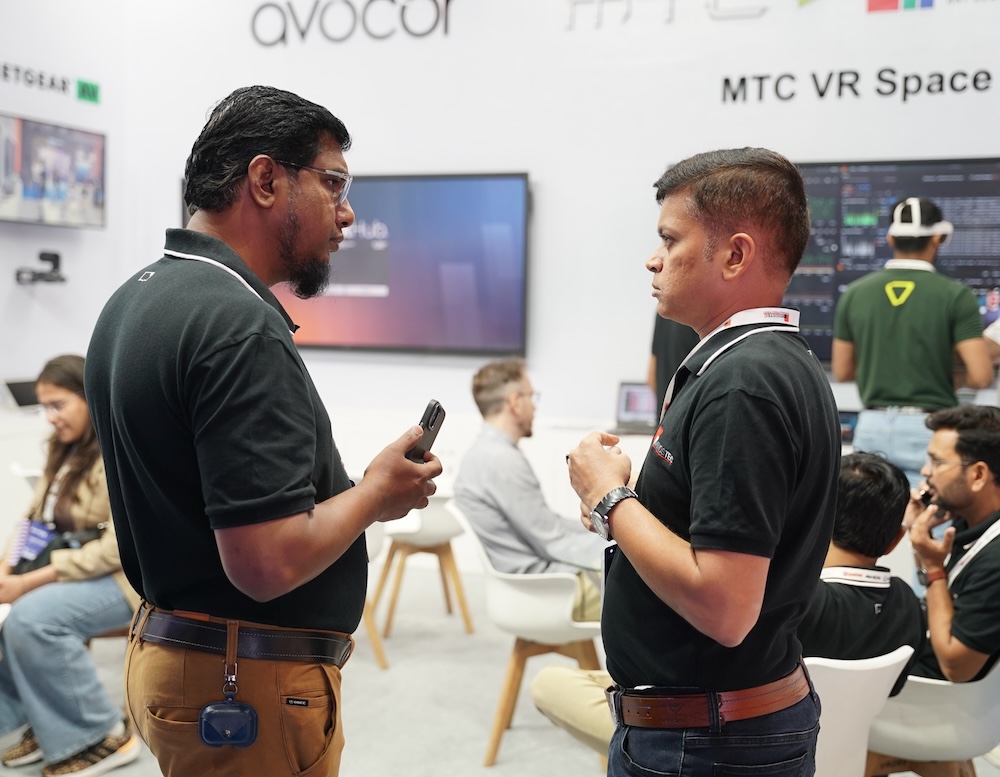 Two men in conversation at a tech exhibition booth labeled "MTC VR Space" with displays and other attendees in the background.