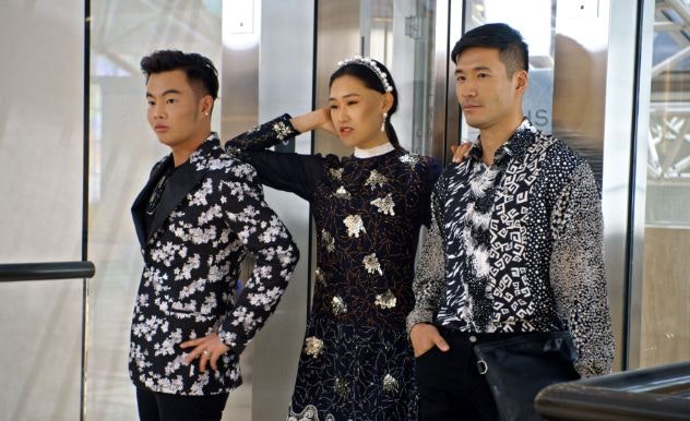 Three people stand in a door way in various fashionable black & white clothing, looking at something off screen