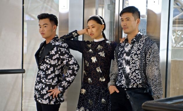 Three people stand in a door way in various fashionable black & white clothing, looking at something off screen