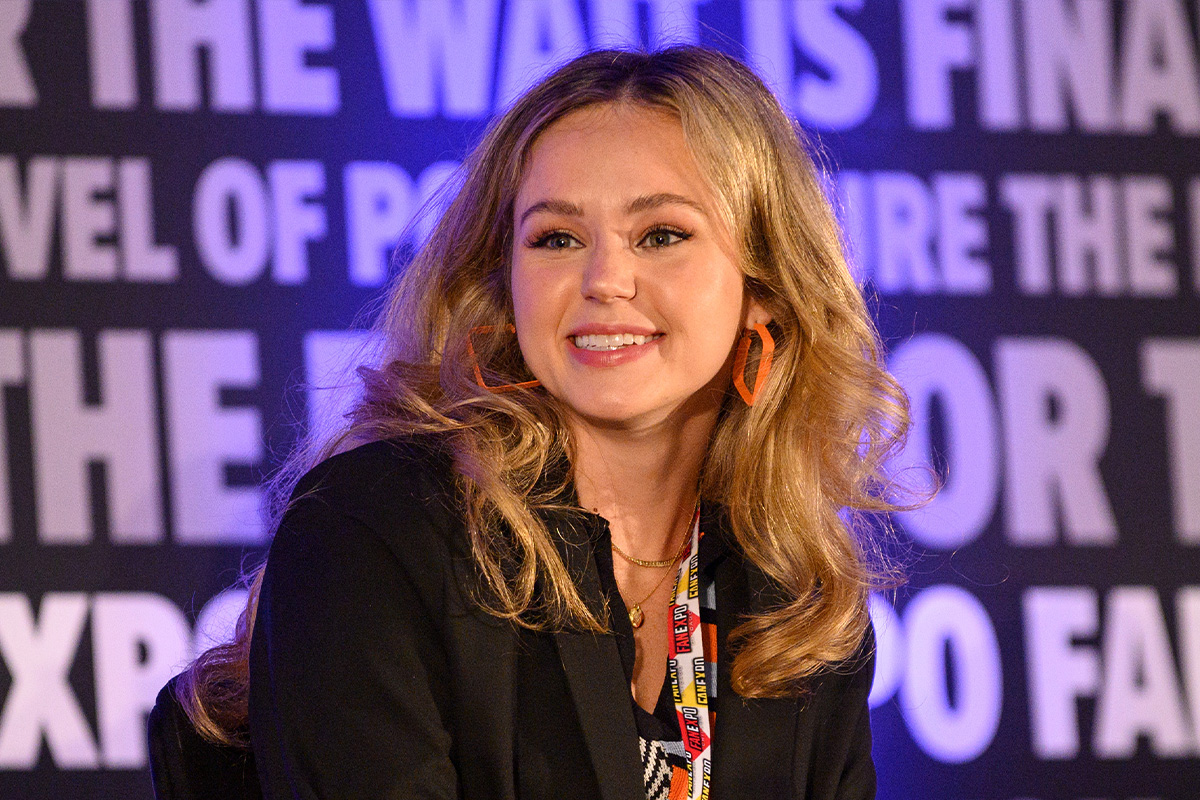 Brec Bassinger head shot. She is smiling and her head is turned slightly to the left. Her blonde hair is down and she is wearing orange square (hoop-like) earrings. She is also wearing a black blazer, yellow/red/white FAN EXPO Lanyard and two gold chains. In the background, is the FAN EXPO background that repeats a variety of phrases.