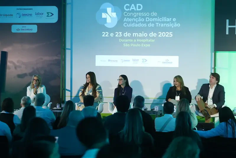 Vista frontal de um painel de discussão no palco do CAD (Congresso de Atenção Domiciliar). Cinco palestrantes estão sentados em poltronas brancas sob um telão que exibe o logotipo do evento e patrocinadores.