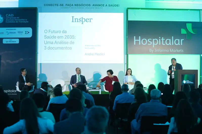 Plano médio do palco durante o Congresso CAD. Ao centro, quatro especialistas estão sentados em um debate. No telão ao fundo, uma apresentação do Insper sobre 'O Futuro da Saúde em 2035' é projetada.