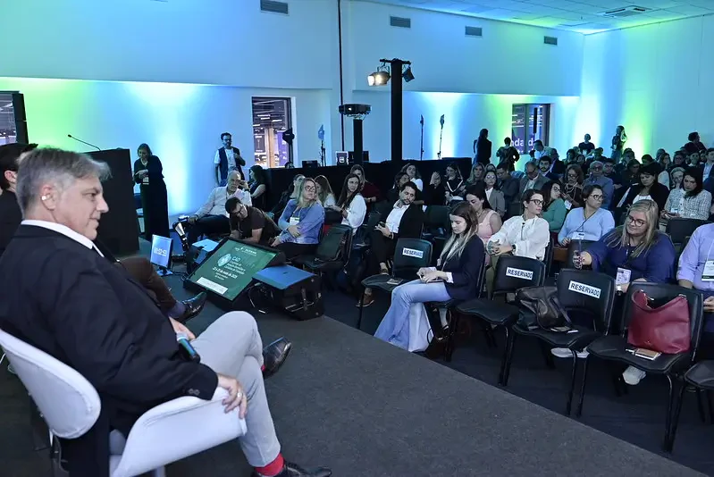 Perspectiva do palco para a plateia. Um palestrante é visto de perfil em primeiro plano, sentado em uma poltrona branca. À sua frente, o público ocupa as cadeiras da sala, com destaque para a iluminação cênica azul nas paredes e o monitor de retorno no chão exibindo o logo do evento.