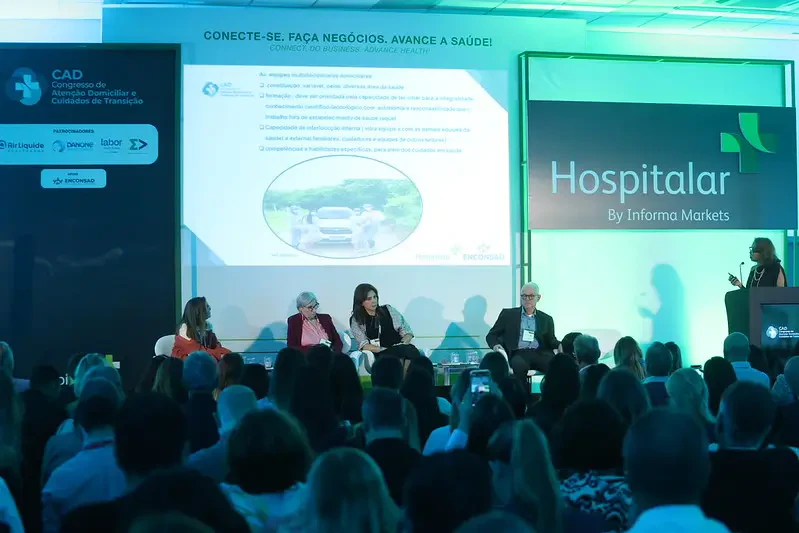 Palestra no Congresso CAD com foco no telão central que detalha informações sobre 'Equipes multidisciplinares domiciliares'. Quatro palestrantes estão sentados no palco e uma mediadora está ao púlpito à direita.