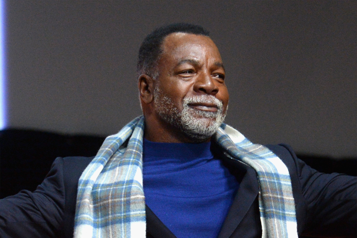Carl Weathers wearing a blue shirt and light blue scarf smiles with his arms extended
