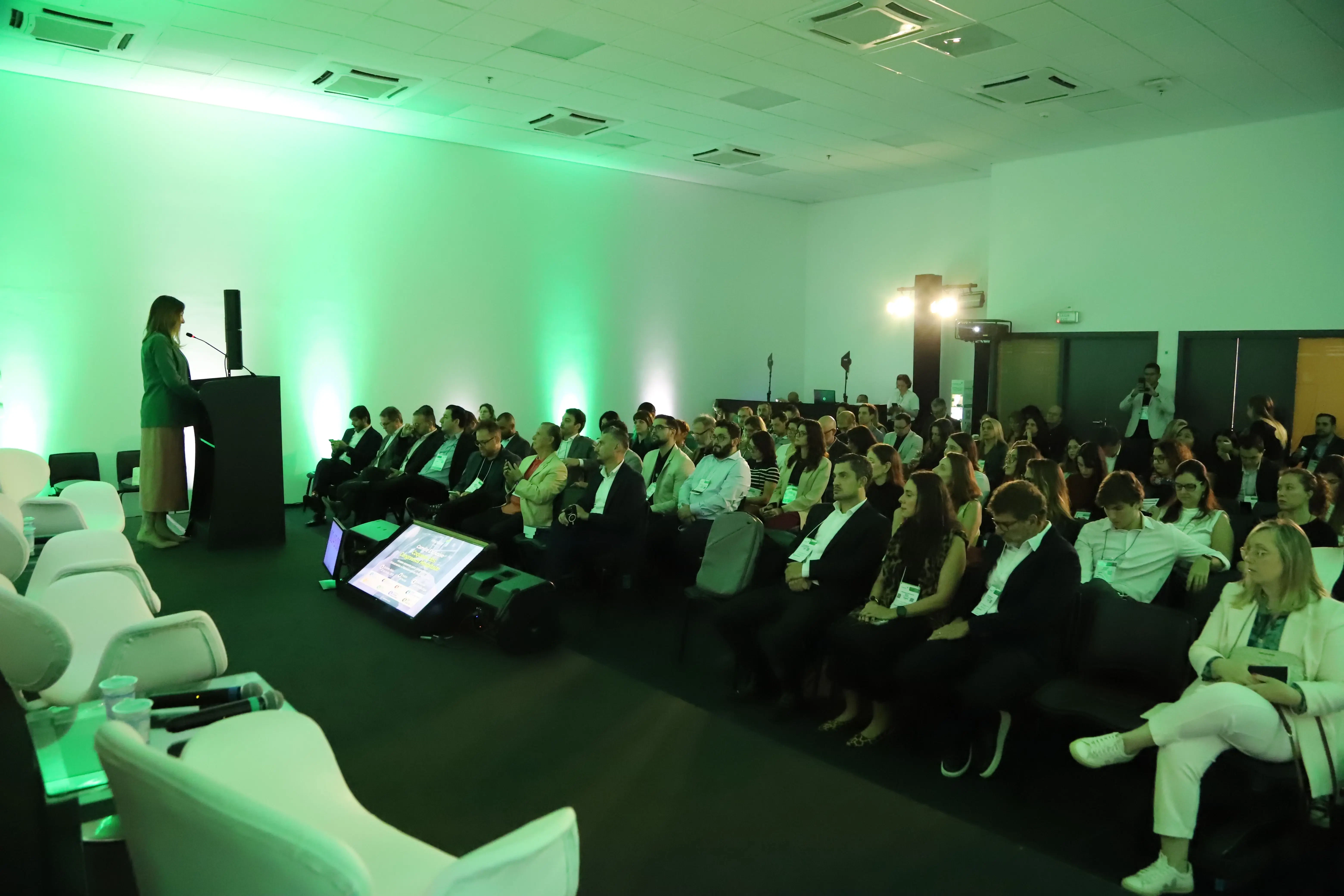 Palestrante feminina em palco com iluminação verde conduzindo conferência corporativa para plateia atenta em auditório moderno. Em destaque, tela de retorno no chão com informações sobre inovação e tecnologia.