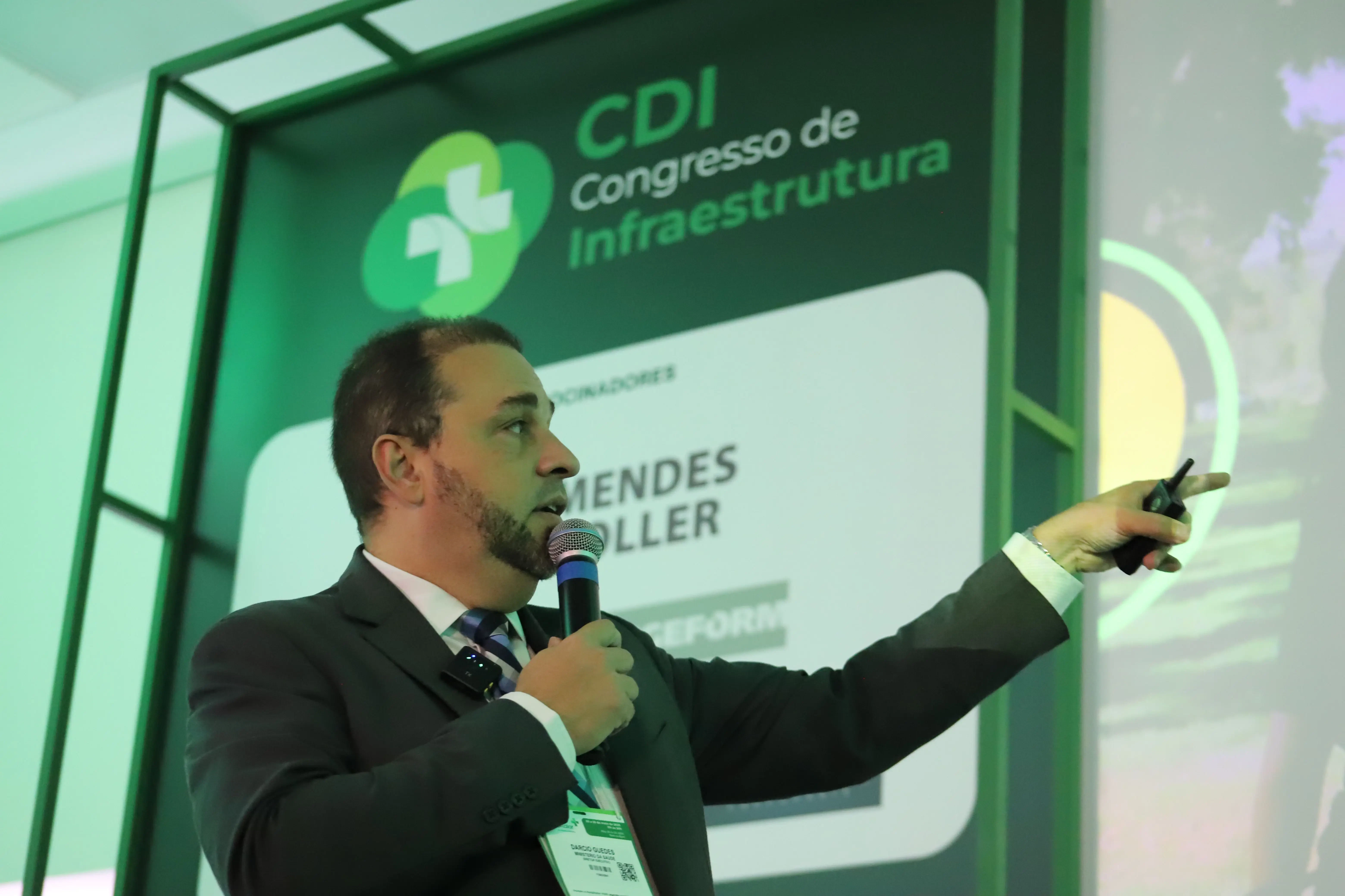 Márcio Guedes, palestrante no CDI Congresso de Infraestrutura, aponta para apresentação enquanto fala ao microfone. No fundo, painel com o logotipo do CDI e patrocinadores como Mendes Holler e Geform.