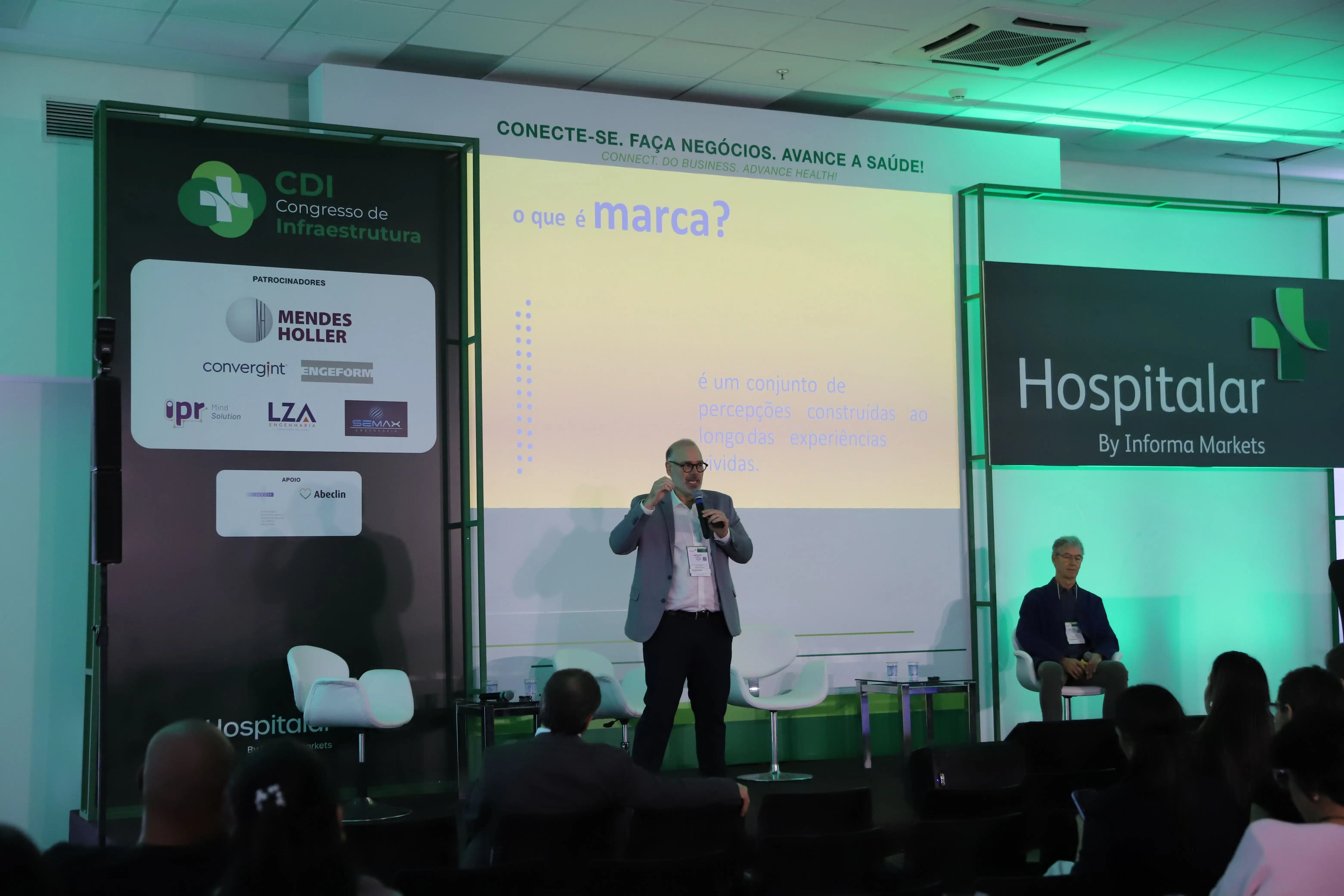 Palestrante no CDI Congresso de Infraestrutura e Hospitalar apresenta slide com a pergunta "O que é marca?" e a definição: "é um conjunto de percepções construídas ao longo das experiências vividas". No palco, logotipos da Hospitalar by Informa Markets e patrocinadores como Mendes Holler e Convergint.