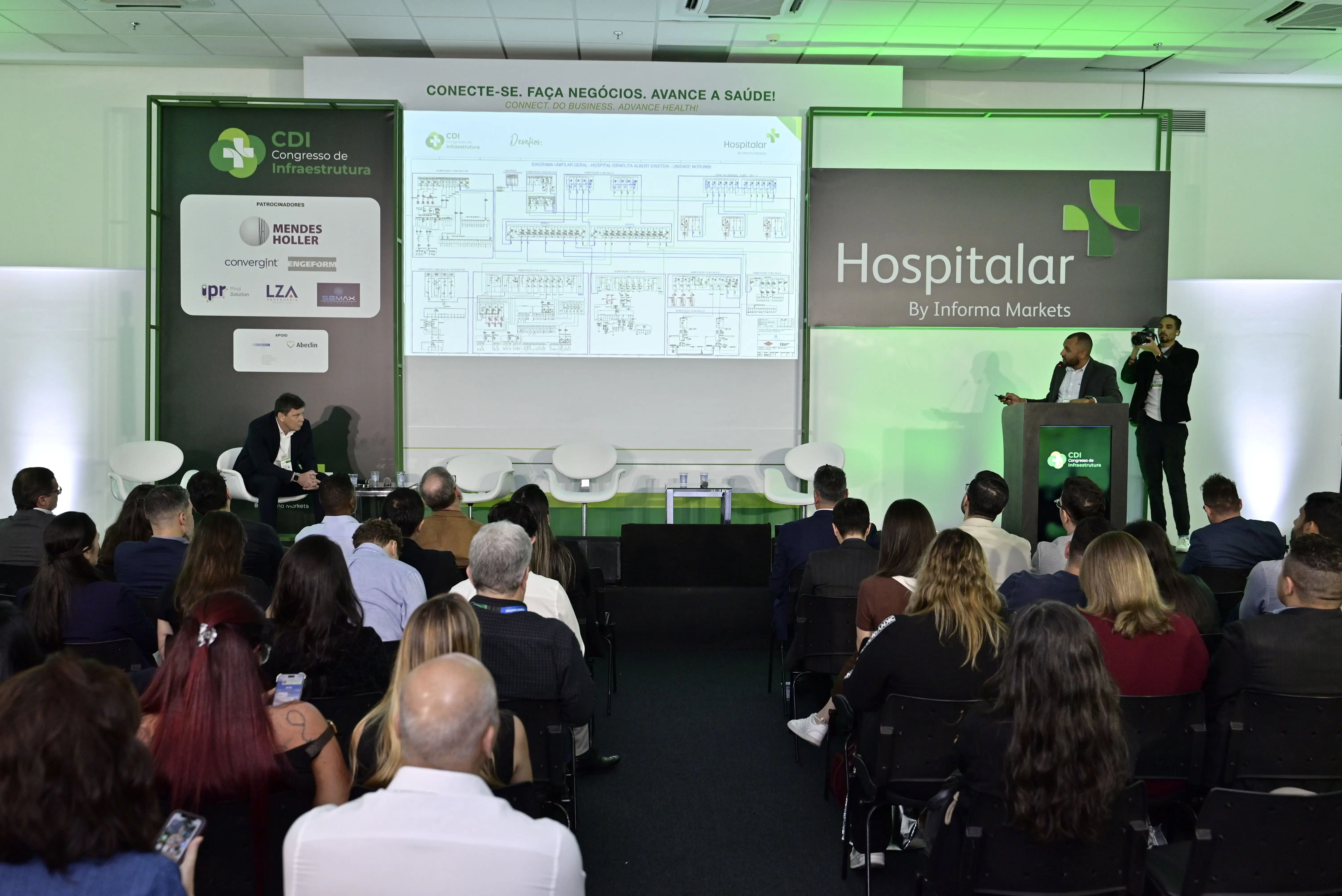 Palestra técnica no CDI Congresso de Infraestrutura e Hospitalar, com palestrante no púlpito apresentando um detalhado diagrama técnico de infraestrutura hospitalar. O palco exibe marcas como Mendes Holler, Convergint e o slogan "Conecte-se. Faça negócios. Avance a saúde!". Plateia atenta ao fundo.
