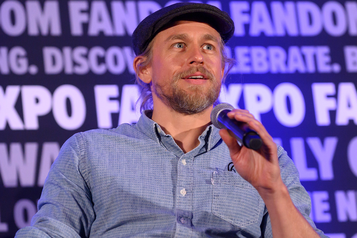A head shot of Charlie Hunnam. There is a microphone tilted up at him. He is half smiling. His hair is pulled back and tucked into the black flat cap. He is wearing a light blue button up with a left  upper pocket with two strings coming out. In the background, is the FAN EXPO background that repeats a variety of phrases.