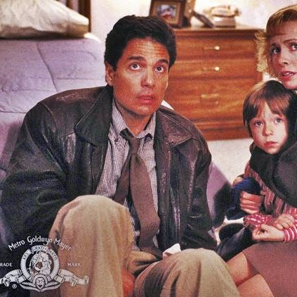 Chris Sarandon wearing a leather jacket, a brown tie, a gray stripped shirt and brown pants. He is sitting on the ground at the end of a bed, looking concerned. Next to him is a woman holding a young child.