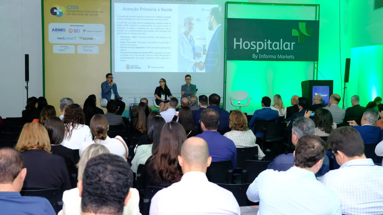 Auditório de evento com plateia sentada assistindo a um painel sobre “Atenção Primária à Saúde”. No palco, três palestrantes participam de uma discussão — um homem fala ao microfone enquanto duas pessoas estão sentadas ao lado. Ao fundo, há um telão com apresentação e, à direita, um painel iluminado em verde com a marca “Hospitalar by Informa Markets”. À esquerda, um banner do CISS (Congresso Internacional de Serviços de Saúde) com logotipos de patrocinadores. O ambiente é corporativo, com iluminação profissional e público atento.
