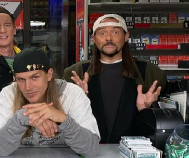Jay and Silent Bob behind a convenience counter. Jay is leaning with his elbows on the counter and is wearing a white shirt over a gray long sleeve hoodie. He is smiling with an eyebrow raised. Silent Bob is next to him with his palms open and his eyes wide open. He is wearing a white cap backwards and dark layer shirts and jacket.
