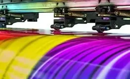 Close dos cabeçotes de impressão de uma impressora plotter industrial passando sobre um vinil de grandes formatos, imprimindo listras vibrantes e embaçadas nas cores magenta, amarelo, preto e ciano (CMYK).