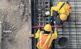 Vista superior de dois operários de construção civil instalando armadura de aço para fundação de concreto, utilizando capacetes amarelos e coletes de segurança refletivos sobre malha de vergalhões.