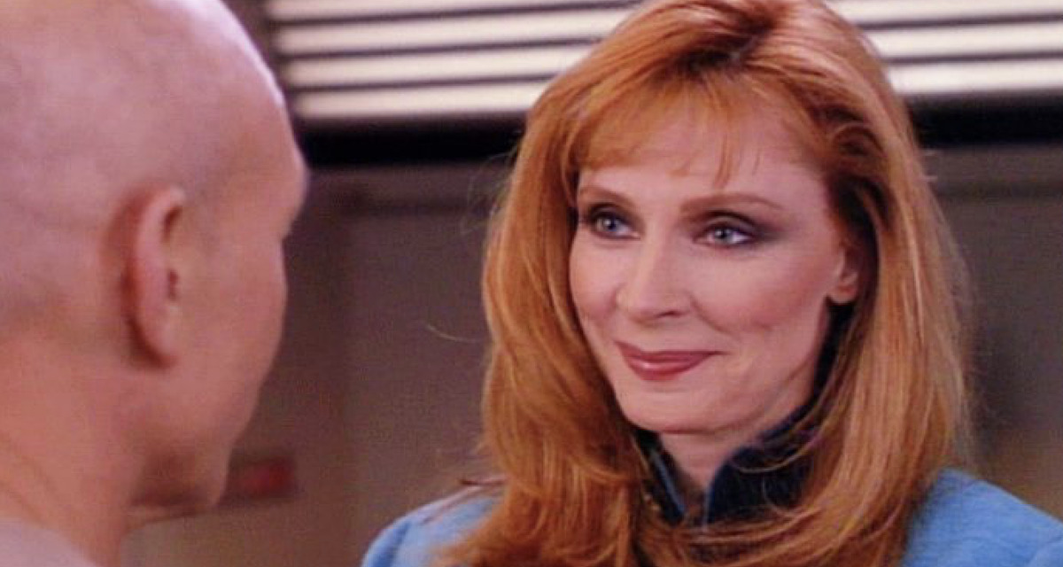 Dr. Crusher smiles, she is in conversation with Jean-Luc Picard, whose head is emerging from the left side of the image.