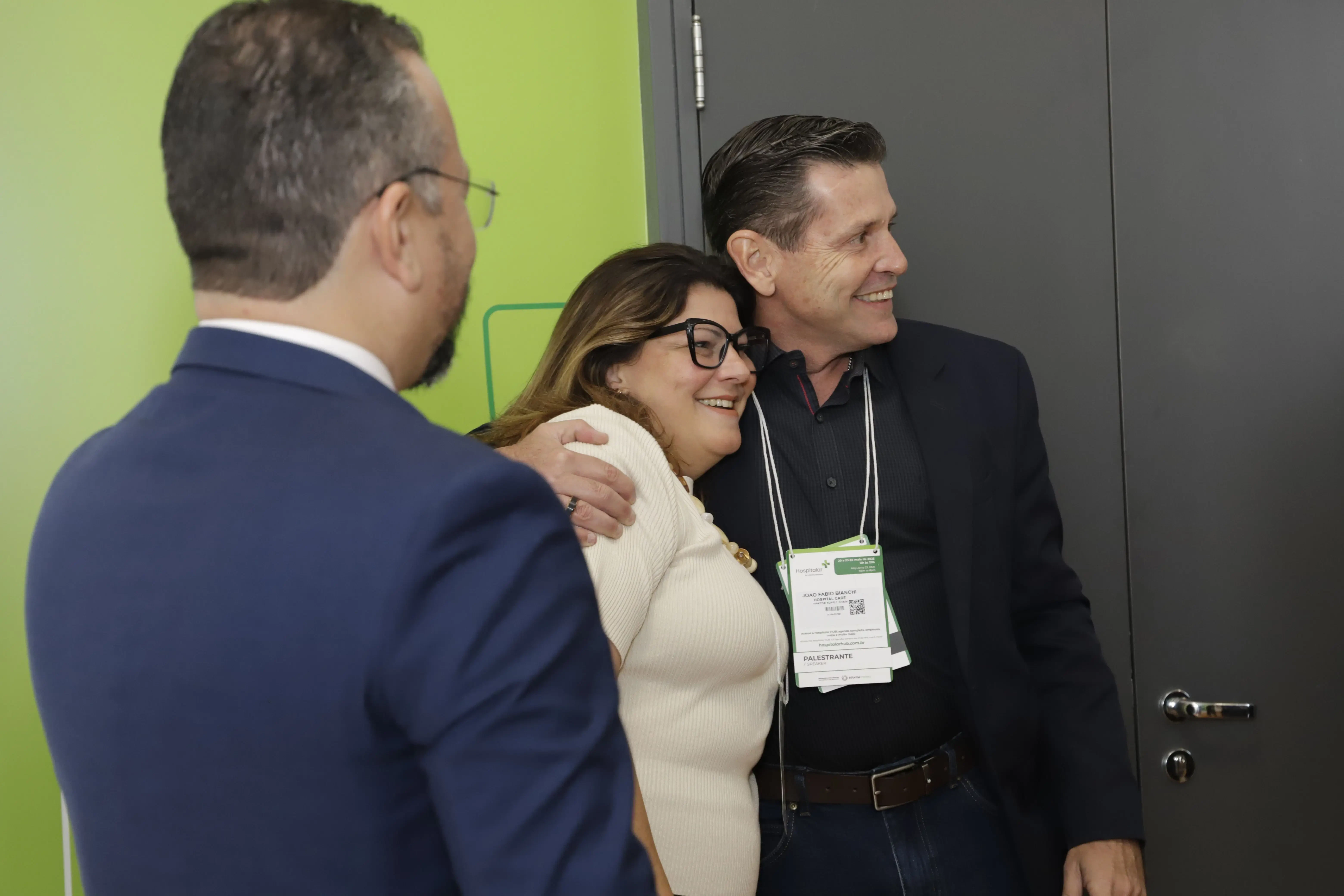 Registro de networking nos bastidores do CSC: Anderson Cremasco (de costas), Ana Paula Melo e outro especialista (João Paulo) sorriem e se abraçam. A imagem reforça o relacionamento entre os curadores e palestrantes do congresso de infraestrutura e saúde.