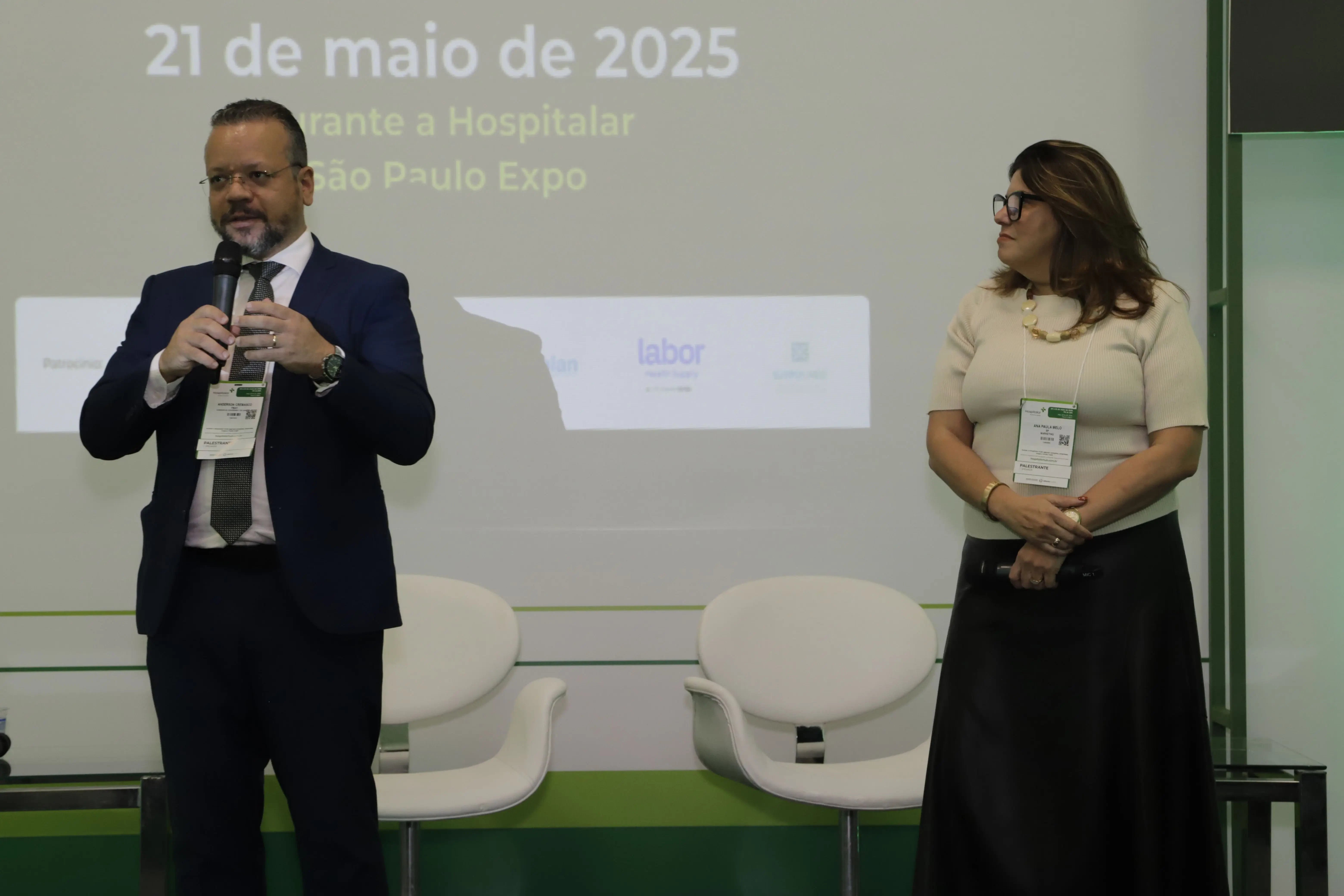 Auditório de evento corporativo com plateia sentada vista por trás. No palco, um homem de terno fala em um púlpito ao lado de um telão que exibe um QR Code e gráficos de inovação. Fundo com iluminação verde. Curadora em pé à direita.