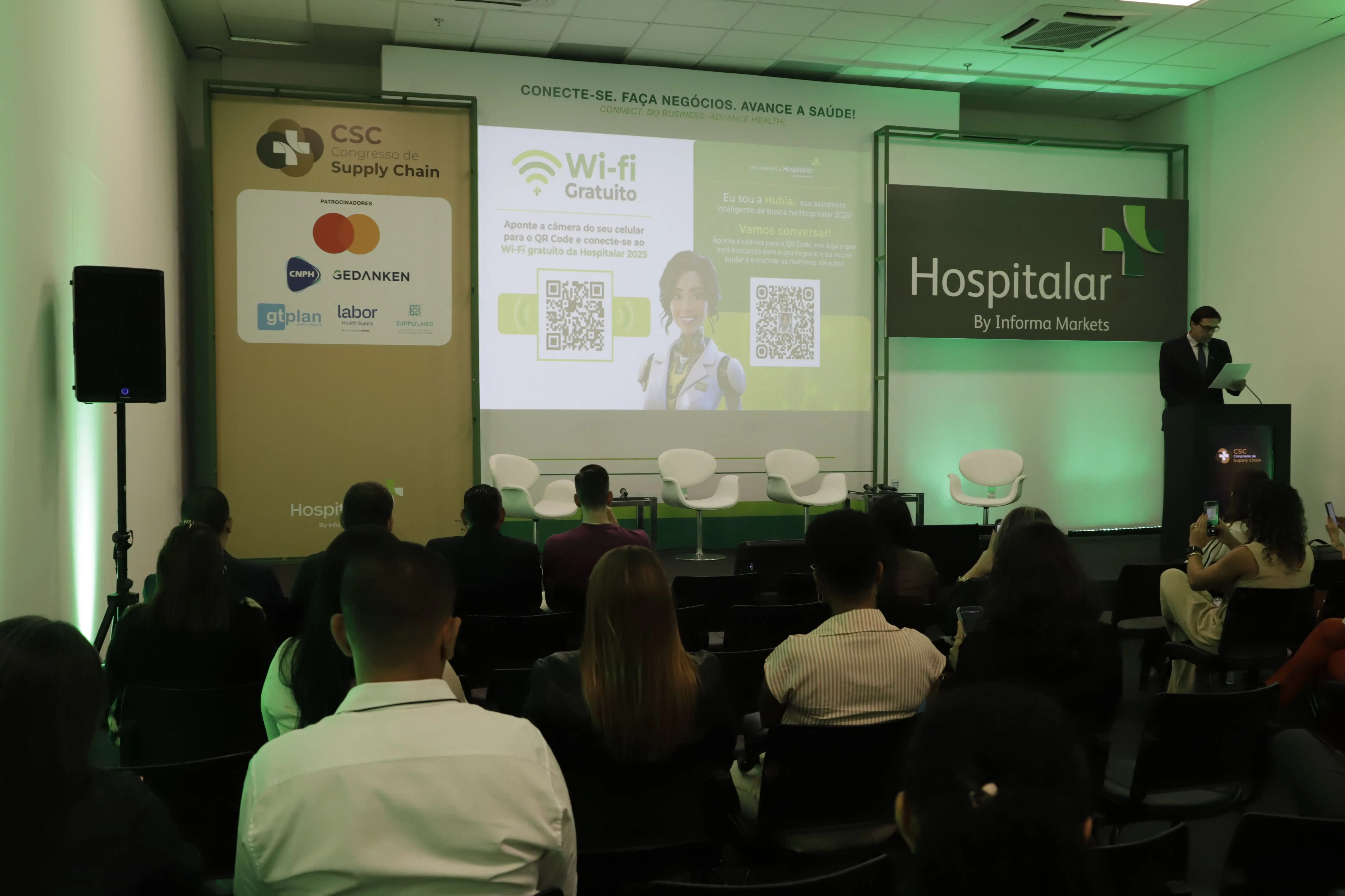 Palestra no CSC Congresso de Supply Chain 2026 sobre "Wi-Fi Gratuito" com uso de QR Code para interação. No palco, apresentador no púlpito ao lado de telão com avatar 3D e logotipos de patrocinadores como Gedanken, GTP Plan e Labor.
