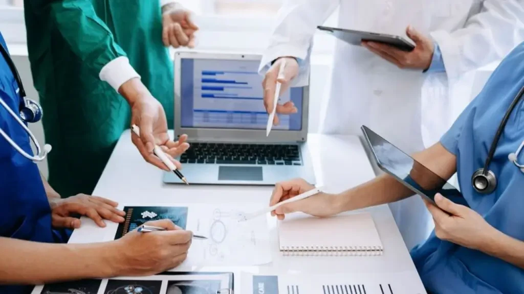 Mãos de equipe médica apontando para gráficos em notebook e relatórios sobre a mesa.