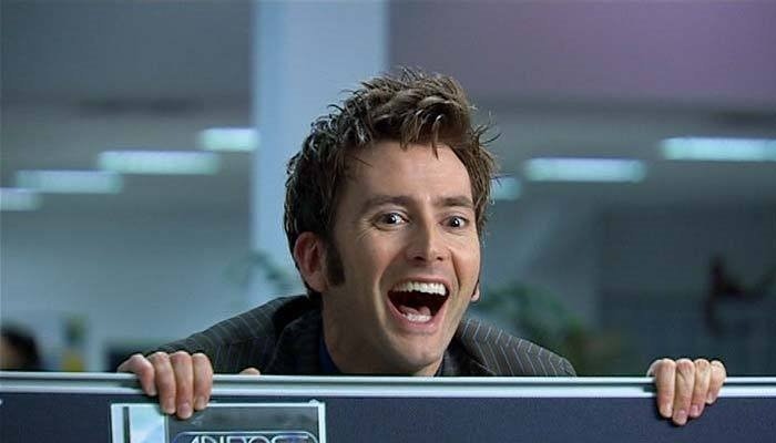 In an office, The Doctor peers over his companion's work cubicle with a joyous, open mouthed smile