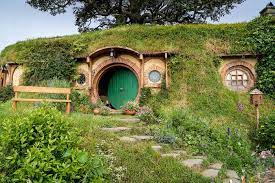 A distant shot of Bag End. The lush grass and greenery encapsulates the whole home - the round green door is ajar.