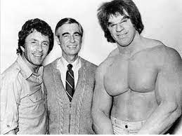 Three men in a black and white photo. Bill Bixby, in a button up shirt, is on the left, Mister Rogers, in his iconic tie and vest is in the middle and Lou Ferrigno, in his hulk costume with no shirt on, is standing on the right.