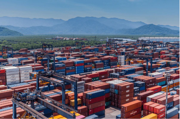 Aerial view of a busy shipping port with stacked cargo containers and cranes, with mountains visible in the background.