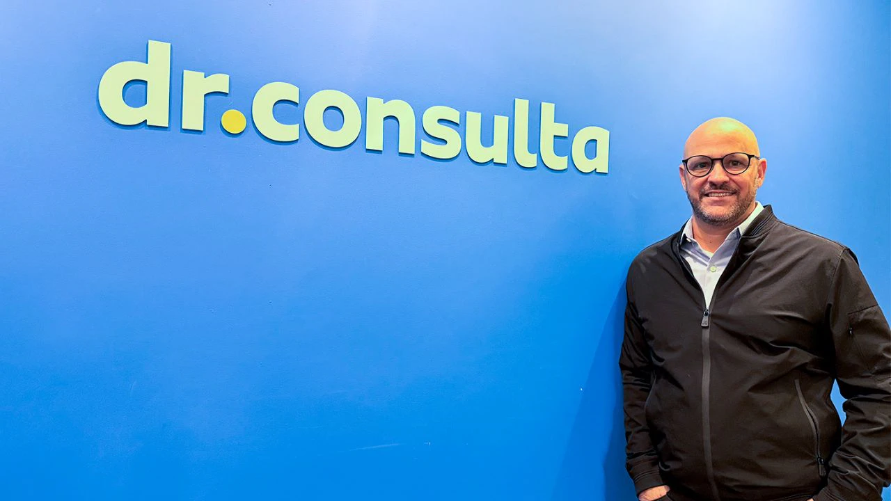 Homem sorridente, usando óculos e jaqueta preta, posa em frente a uma parede azul com o logotipo “dr.consulta” em destaque. Ele está de pé, com postura relaxada, em um ambiente corporativo limpo e moderno.
