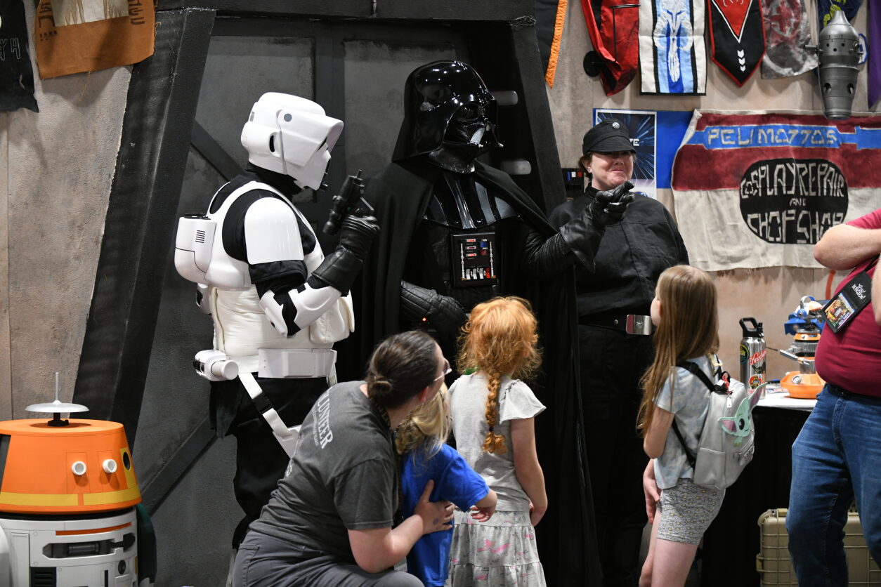 Children clamor around Darth Vader and his guard, staring in awe. A Storm trooper stands at one side, and imperial guard on the other.