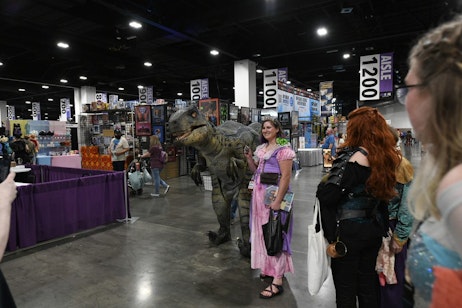 An eager fan, dressed as Rapunzel, poses with Onyx the Velociraptor on the show floor, as a line of fans wait to get their photo as well