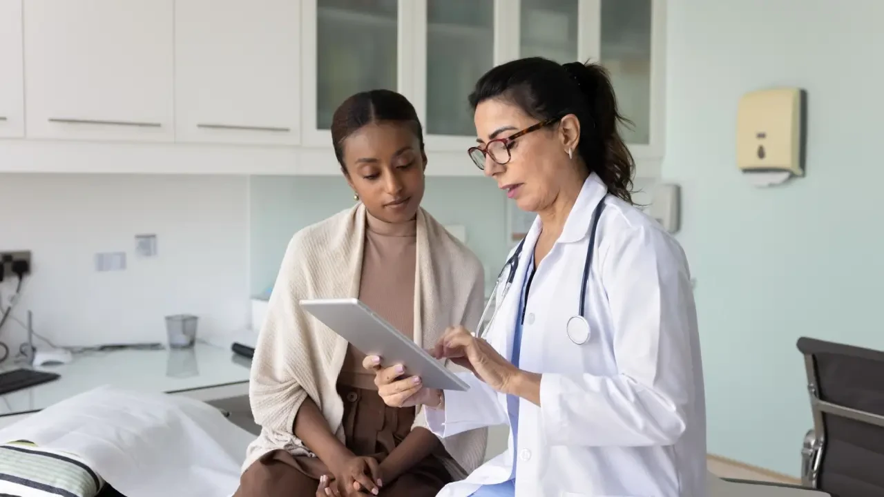 Médica em jaleco branco utiliza um tablet para explicar resultados de exames a uma paciente em consultório moderno. A imagem ilustra o uso de tecnologia e saúde digital no atendimento ao paciente, refletindo as inovações em gestão hospitalar apresentadas na Hospitalar 2026.