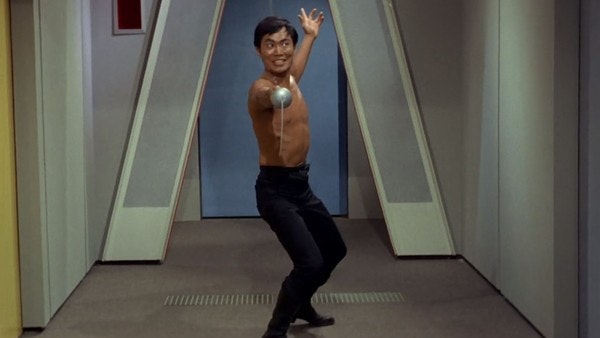 Sulu stands shirtless in the hall of the Starship Enterprise. He is in starting position for a joust, with the sword pointing straight towards the camera.