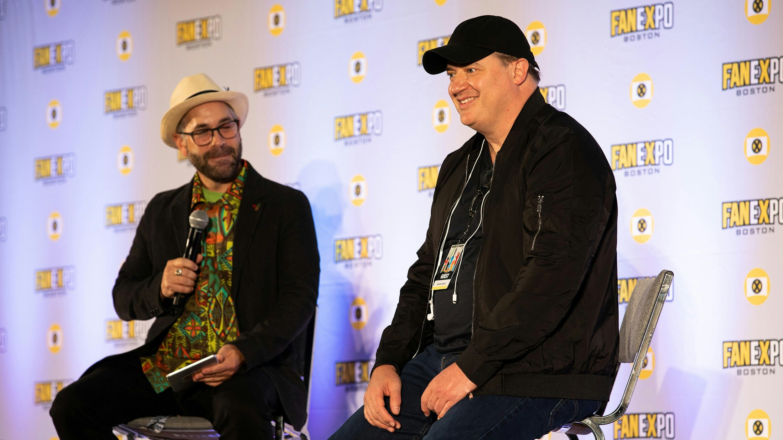 Brendan Fraser sitting in a black jacket with a black hat smiling on stage
