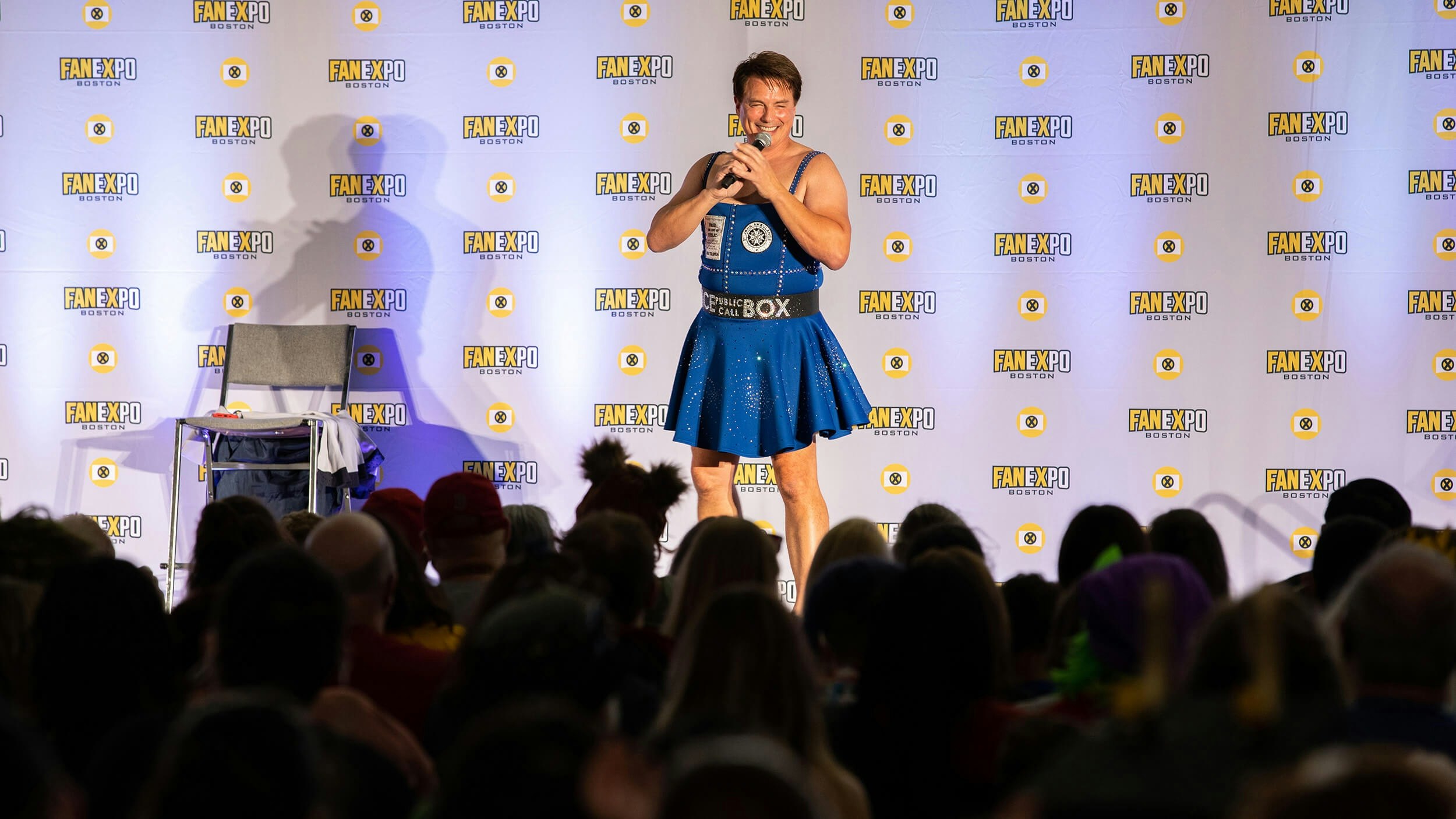 John Barrowman smiles while holding a microphone in a blue TARDIS dress
